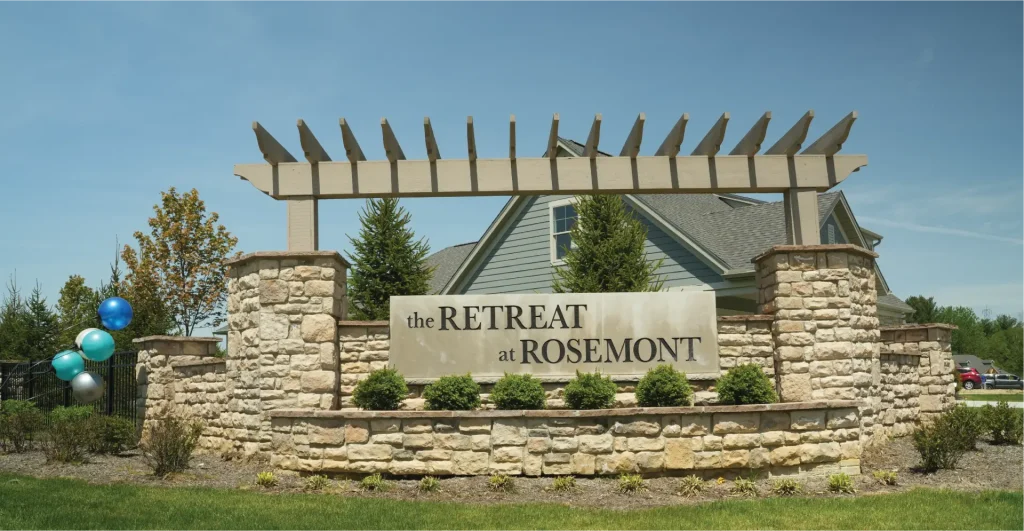 A sign for "The Retreat at Rosemont" displayed on a stone wall under a wooden pergola, with a house and trees visible in the background.